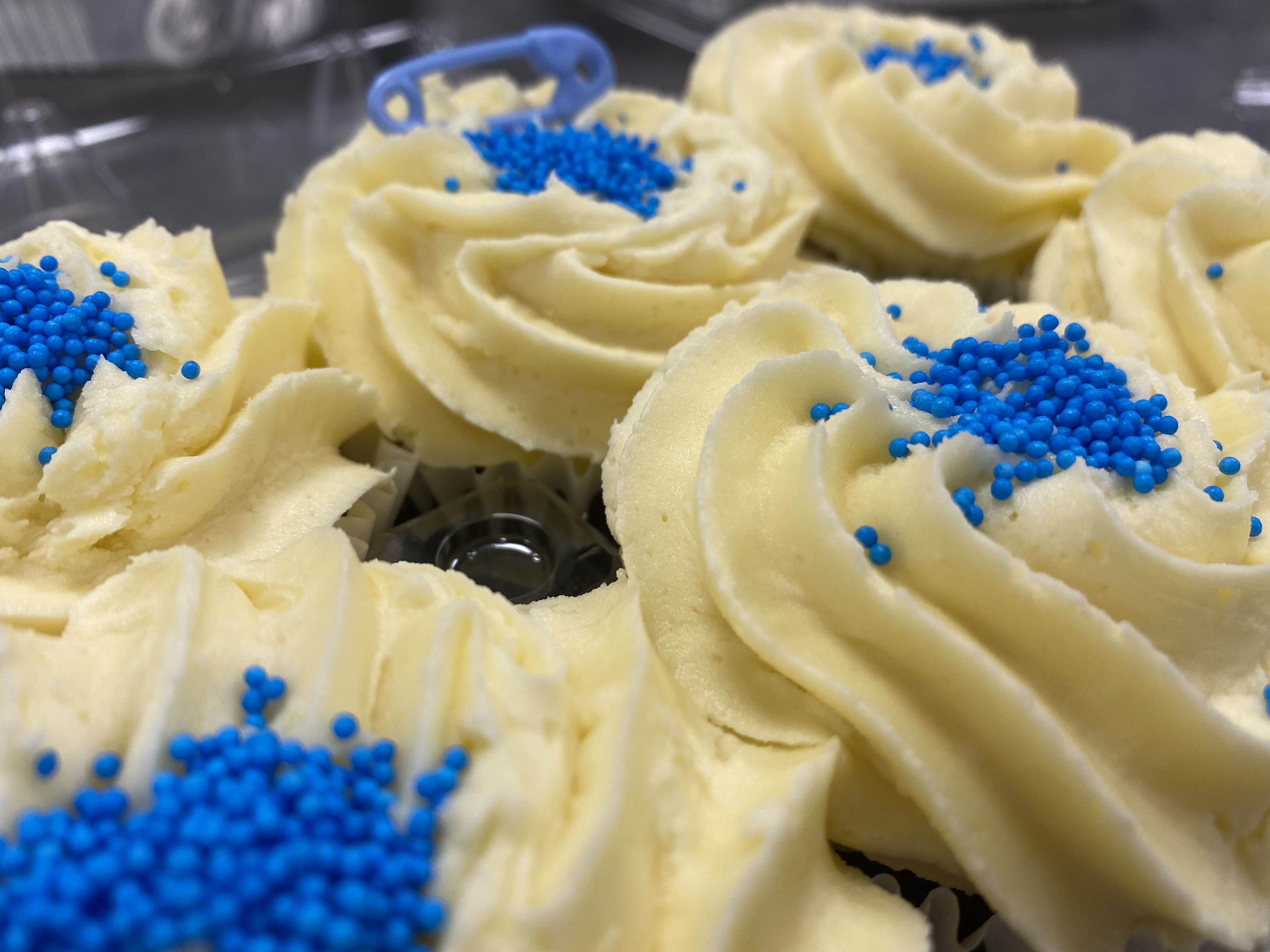 Close-up image of vanilla cupcakes with vanilla buttercream frosting, topped with blue sprinkles. These delightful treats are packed together in a plastic container, and a small blue decoration can be seen on one, reminiscent of charming wedding cakes.